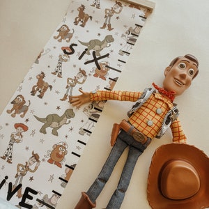 May include: A white and black growth chart with a Toy Story theme. The chart features a variety of Toy Story characters, including Woody, Buzz Lightyear, and Rex. The chart is marked with numbers from one to six. A Woody doll is lying on the floor next to the chart.