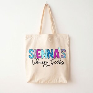 May include: A beige tote bag with the text "SIENNA'S Library Books" in colorful letters. The letters are decorated with a patchwork design.