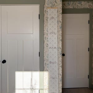 May include: A white wooden growth chart with black numbers and floral illustrations on a white background. The chart is mounted on a wall with green floral wallpaper.