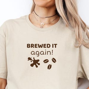 May include: A beige t-shirt featuring the text "BREWED IT again!" in brown, accompanied by coffee bean and spill graphics. The shirt is worn with blue jeans.
