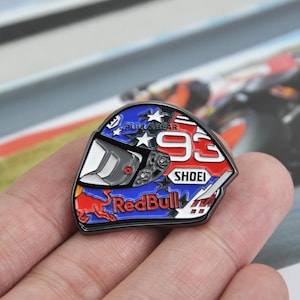 May include: A motorcycle helmet badge in blue, red, and white. The badge features the number '93', the 'Red Bull' logo, and the 'Shoei' brand. The helmet has a visor and stars.