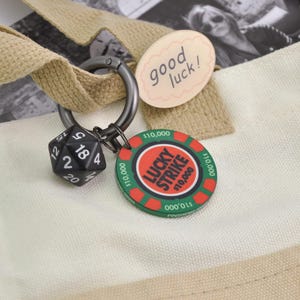 May include: A black 20-sided die and a Lucky Strike poker chip are attached to a metal carabiner. The poker chip is red and green with the text "LUCKY STRIKE $10,000". A tag with the words "good luck!" is also attached.