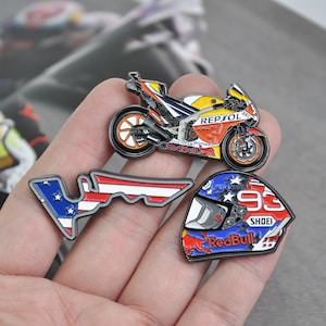 May include: A collection of motorcycle-themed enamel pins. The pins include a detailed motorcycle, a helmet with the number 93, and a racetrack outline with an American flag design. The pins feature the Red Bull and Repsol logos.