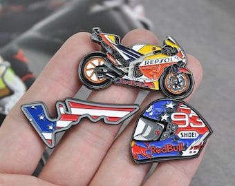 May include: A collection of motorcycle-themed enamel pins. The pins include a detailed motorcycle, a helmet with the number 93, and a racetrack outline with an American flag design. The pins feature the Red Bull and Repsol logos.