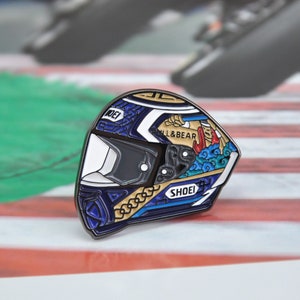 Puede incluir: Pin de casco de moto con diseño detallado. El pin presenta una visera blanca, detalles en azul y dorado, y el texto "SHOEI" y "PULL&BEAR". El pin se encuentra sobre un fondo borroso de un circuito.