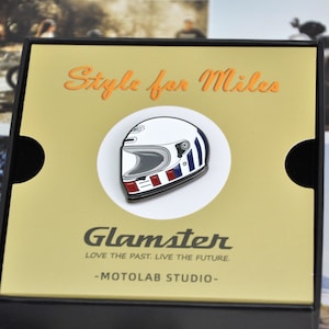 Pode incluir: Um alfinete de capacete de motocicleta com base branca, listras azuis e vermelhas e viseira prateada. O alfinete está sobre um fundo dourado com as palavras "Style for Miles" em laranja. A marca "Glamster" e "-MOTOLAB STUDIO-" também são visíveis.