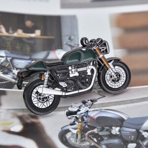Puede incluir: Un pin de esmalte detallado de una motocicleta clásica en verde oscuro y negro. El pin presenta detalles intrincados del motor, las ruedas y el marco, con detalles dorados en las horquillas delanteras. La motocicleta está sobre un fondo borroso.