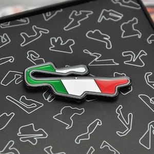 Könnte beinhalten: Ein Ansteck-Pin mit einem Rennstrecken-Design in den Farben der italienischen Flagge: Grün, Weiß und Rot. Der Pin befindet sich vor einem schwarzen Hintergrund mit weißen Rennstrecken-Umrissen.