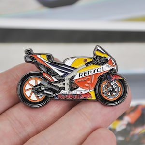 May include: A motorcycle pin with a detailed design. The pin features a yellow, orange, white, and blue motorcycle with the words "REPSOL" and "Red Bull" visible. The pin is held in a person's hand.