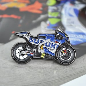May include: A blue and white Suzuki motorcycle pin with the word "SUZUKI" prominently displayed. The pin features black wheels, a detailed engine, and the "Eckstar" logo. The pin is a collectible item.