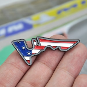 Può includere: Una spilla di metallo a forma di circuito, con il design della bandiera americana. La spilla ha un bordo grigio, con una sezione blu contenente stelle bianche e strisce rosse e bianche. La spilla è tenuta in mano.