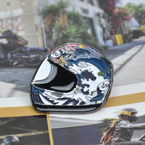 May include: A motorcycle helmet pin with a Japanese wave design in blue, white, and black. The pin features floral accents and the word "Arai". The helmet is a motorcycle accessory.
