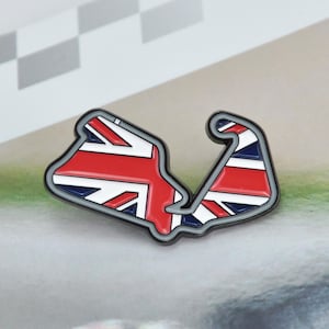 Puede incluir: Un pin metálico con un diseño de circuito de carreras que incorpora la bandera del Reino Unido. El pin presenta los colores rojo, blanco y azul dentro de un contorno gris. El diseño es una representación estilizada del Reino Unido.