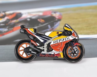 May include: A detailed enamel pin of a Repsol Honda RC213V motorcycle. The pin features a yellow, orange, white, and blue color scheme with black wheels and accents. The pin has the Red Bull and Repsol logos.