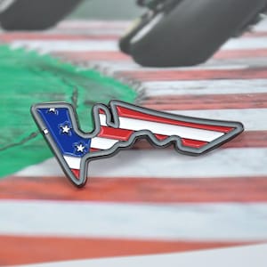 May include: A pin shaped like a race track, featuring the American flag design. The pin has a blue section with white stars and red and white stripes. It has a gray border and is likely a collectible item.
