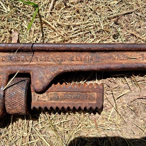 May include: Close-up of a rusty, vintage pipe wrench. The wrench is brown with visible text that reads "THE RIDGE TOOL CO. ELYRIA" and "HARDENED 14". The tool is lying on a bed of dry grass.