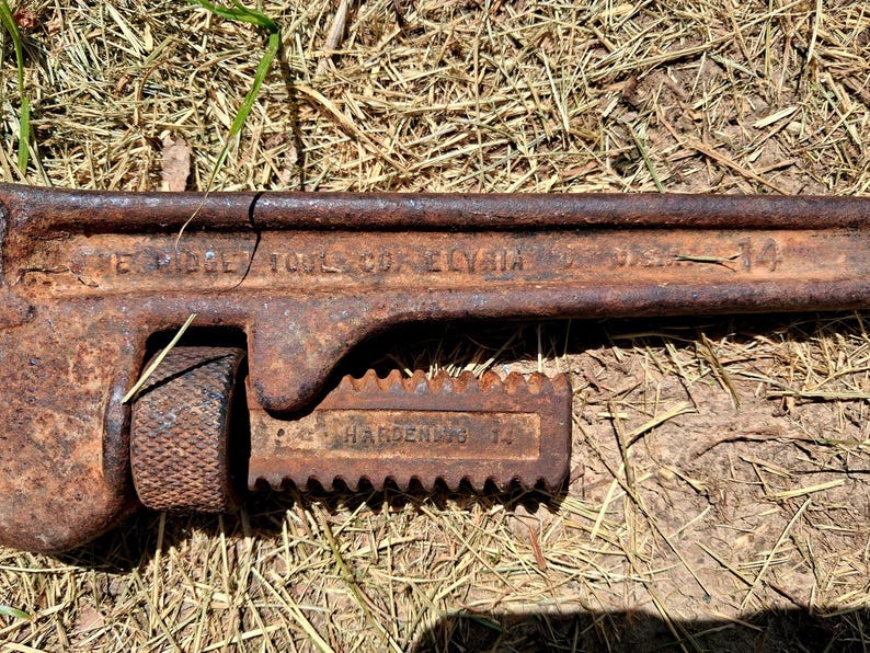 May include: Close-up of a rusty, vintage pipe wrench. The wrench is brown with visible text that reads "THE RIDGE TOOL CO. ELYRIA" and "HARDENED 14". The tool is lying on a bed of dry grass.