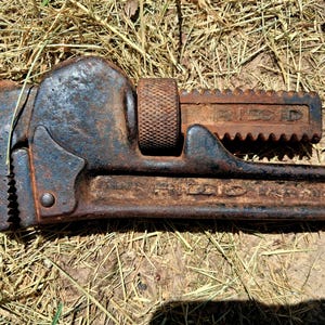 May include: A vintage, rusty pipe wrench with the brand name "RIDGID" visible. The wrench is made of dark metal with signs of wear and tear, including rust. The adjustable jaw is open, and the tool lies on a bed of dry grass.