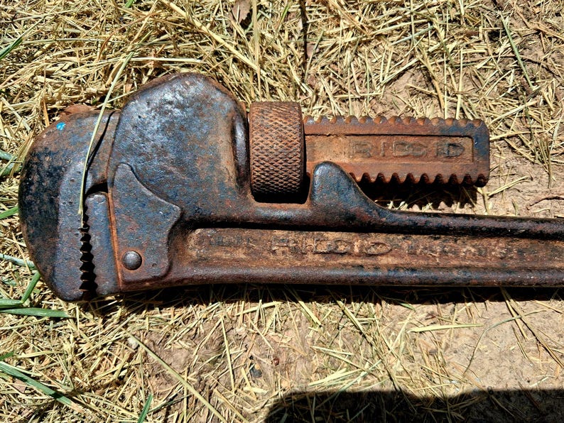 May include: A vintage, rusty pipe wrench with the brand name "RIDGID" visible. The wrench is made of dark metal with signs of wear and tear, including rust. The adjustable jaw is open, and the tool lies on a bed of dry grass.