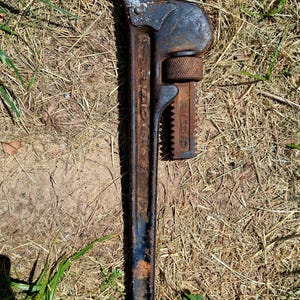 May include: A vintage, rusty pipe wrench, lying on dry grass and dirt. The wrench is dark blue and brown with visible wear and tear. The brand name "RIDGID" is visible on the side of the wrench.