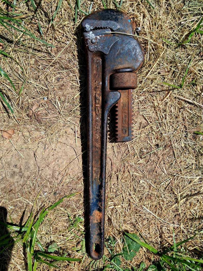 May include: A vintage, rusty pipe wrench, lying on dry grass and dirt. The wrench is dark blue and brown with visible wear and tear. The brand name "RIDGID" is visible on the side of the wrench.