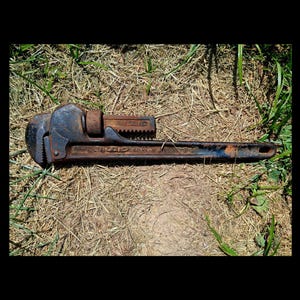 May include: A vintage, rusty pipe wrench with a dark blue and brown finish. The wrench is lying on a bed of dry grass and green blades. The adjustable jaws and handle are clearly visible, showcasing its industrial design.