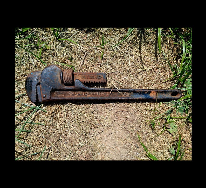 May include: A vintage, rusty pipe wrench with a dark blue and brown finish. The wrench is lying on a bed of dry grass and green blades. The adjustable jaws and handle are clearly visible, showcasing its industrial design.