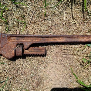May include: A rusty, vintage pipe wrench, lying on dry grass. The wrench is a dark brown color, with visible rust and wear. It has a long handle and an adjustable jaw, typical of this type of tool. The wrench is a size 14.