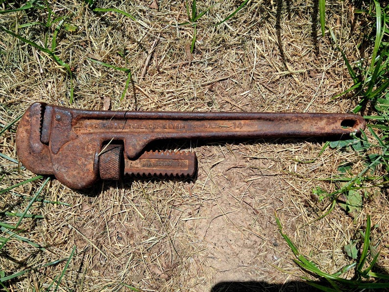 May include: A rusty, vintage pipe wrench, lying on dry grass. The wrench is a dark brown color, with visible rust and wear. It has a long handle and an adjustable jaw, typical of this type of tool. The wrench is a size 14.