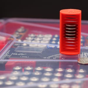 May include: A red plastic battery holder with a clear plastic case. The holder has multiple slots for coin-shaped batteries. The text "Battery Pack" is visible on the case.