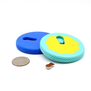 Puede incluir: Dos tapones de botella de silicona coloridos, uno azul y otro turquesa con un acento amarillo, están junto a una moneda y un pequeño objeto marrón. Los tapones tienen un borde texturizado y una ranura.