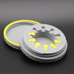 Op de afbeelding: Een grijze plastic container met een gele rand en een grijs deksel. Het deksel heeft een cirkelvormig patroon van gele lipjes rond de rand.