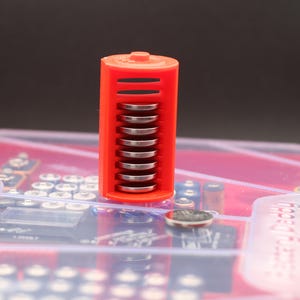 Puede incluir: Un soporte de baterías de plástico rojo con una caja de plástico transparente. El soporte está lleno de una pila de baterías redondas plateadas.