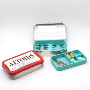 May include: Three small metal and plastic containers. One is a red and silver Altoids tin. Another is a silver tin with a teal interior. The third is a teal plastic container with compartments, holding pills and keys.