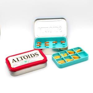 May include: An open silver Altoids tin with a turquoise interior and orange mint holders, next to a turquoise mint tray filled with yellow mints. A closed red and silver Altoids tin with white text is also present.