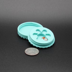 May include: Two turquoise plastic containers with a paw print design. The top container has a small round opening with a silver button inside.