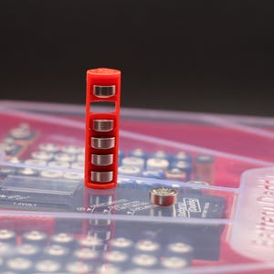 Puede incluir: Un contenedor de plástico rojo con cinco pilas de botón plateadas en su interior. El contenedor está de pie sobre una superficie de plástico transparente con un fondo rojo. El texto "Battery Daddy" es visible en el fondo.