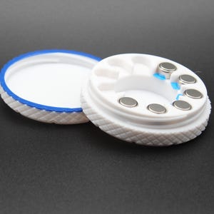 May include: A white round container with a blue rim, containing small round silver magnets. The container is open and the lid is next to it.