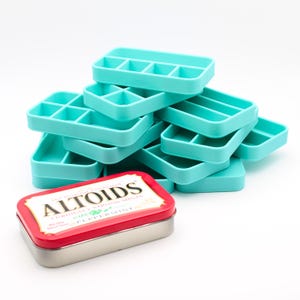 May include: A stack of turquoise plastic organizers with multiple compartments, next to a red and silver Altoids tin. The tin has the words "Altoids Curiously Strong Mints Peppermint" printed on it.