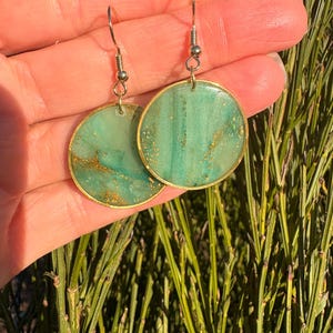 May include: A pair of round earrings with a teal and gold marbled design. The earrings have a gold-colored metal frame and ear hooks. The earrings are held in a person's hand against a background of green foliage.
