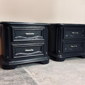 May include: Two matching black nightstands with two drawers each. The nightstands have ornate detailing and silver-toned hardware. The drawers have a decorative wave design. The nightstands are on a gray and white speckled floor.