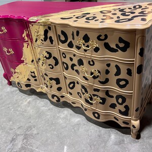 May include: A unique dresser with a two-tone design. One side is magenta, the other is tan with black leopard spots. Gold-colored hardware and trim add a touch of elegance. The dresser has three drawers and a curved front.