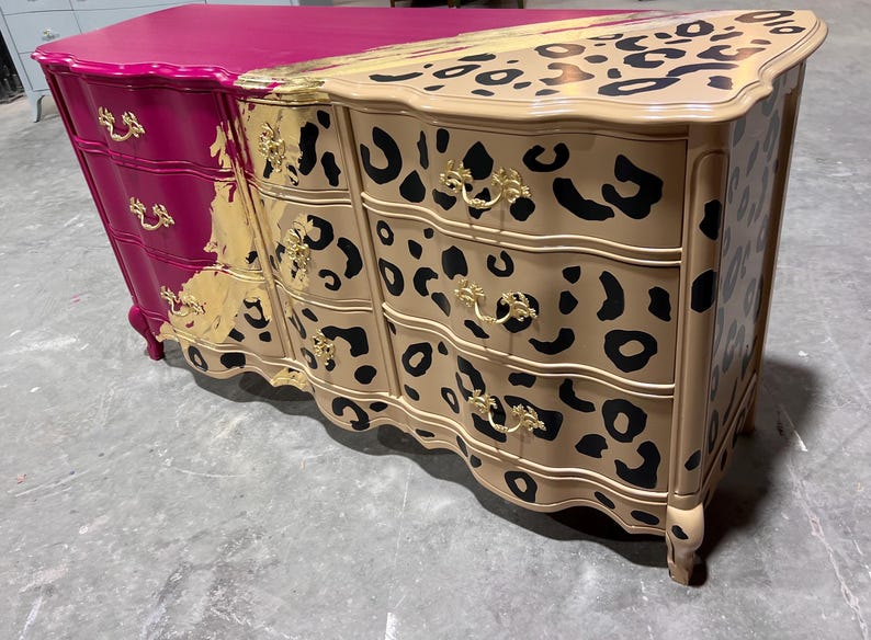 May include: A unique dresser with a two-tone design. One side is magenta, the other is tan with black leopard spots. Gold-colored hardware and trim add a touch of elegance. The dresser has three drawers and a curved front.