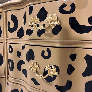 May include: A tan dresser with black leopard spots and gold-colored ornate drawer pulls. The dresser has a curved design and a glossy finish, adding a touch of elegance to the furniture piece.
