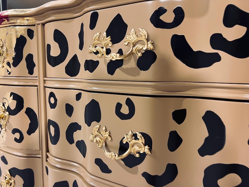 May include: A tan dresser with black leopard spots and gold-colored ornate drawer pulls. The dresser has a curved design and a glossy finish, adding a touch of elegance to the furniture piece.