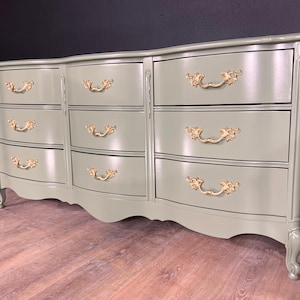 May include: A sage green dresser with nine drawers and ornate gold-colored handles. The dresser has a curved front and decorative legs, adding a touch of elegance to the bedroom furniture.