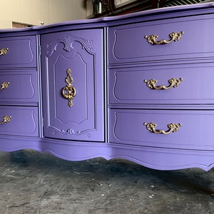 May include: A lavender dresser with ornate gold hardware. The dresser has seven drawers and a cabinet door. The drawers and cabinet door have decorative molding. The dresser has curved legs and a curved bottom edge.