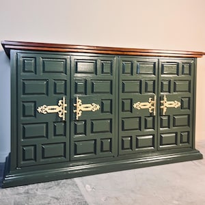 May include: A dark green cabinet with four doors and decorative gold-colored hardware. The cabinet has a dark wood top and a rectangular design on the doors. The cabinet is a piece of furniture.