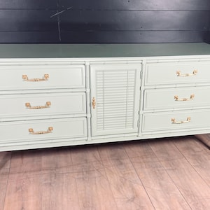 May include: A light green dresser with six drawers and a center cabinet with a slatted door. The dresser has gold-colored handles and sits on four legs. The top of the dresser is a darker green.