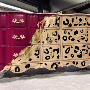 May include: A painted dresser with a unique design. The left side is a deep magenta, while the right side features a tan background with black leopard spots. Gold leaf accents and ornate hardware complete the look. The dresser has six drawers.